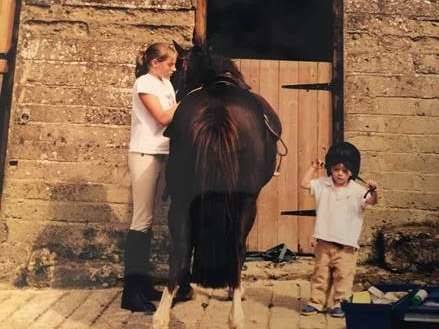 One girl, a little boy and a black horse in a stable.